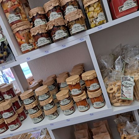 Ein weißer Regalboden mit verschiedenen Gläsern gefüllt mit konservierten Früchten und Gemüse, gekennzeichnet mit 'Chironi'. Die Gläser sind in verschiedenen Größen und Farben. Unter dem Regal befinden sich Packungen von Brot und anderen Snacks.