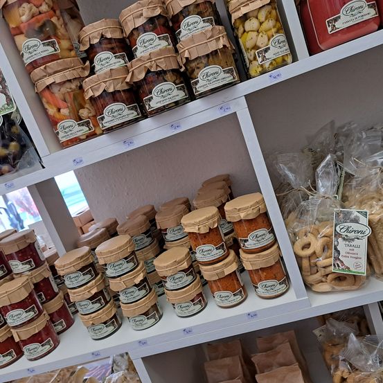 Ein weißer Regalboden mit verschiedenen Gläsern gefüllt mit konservierten Früchten und Gemüse, gekennzeichnet mit 'Chironi'. Die Gläser sind in verschiedenen Größen und Farben. Unter dem Regal befinden sich Packungen von Brot und anderen Snacks.