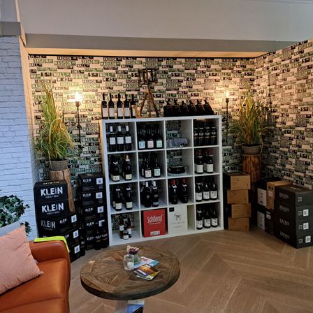 Ein Innenraum mit einem Holztisch, einem weißen Regal voller Weinflaschen und Wänden, die mit Straßenschildern geschmückt sind. Ein Topf mit Pflanzen steht neben dem Regal.