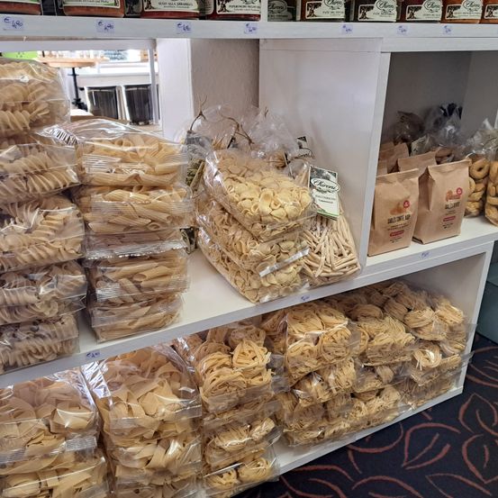 Ein Ladenregal zeigt verschiedene Nudelarten in Plastiktüten und Papiertüten. Einige Nudeln sind spiralförmig, andere dünn und wieder andere in verschiedenen Formen. Die Nudeln sind ordentlich auf den Regalen angeordnet.