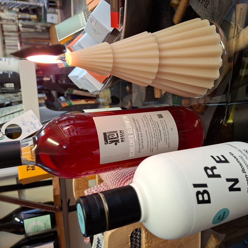 Eine Flasche Rotwein steht auf einem Tisch mit anderen Gegenständen. Die Flasche ist mit einem weißen Etikett versehen, und eine weitere weiße Flasche mit der Aufschrift 'BIRE' befindet sich in der Nähe. Ein Glasbehälter mit einer brennenden Kerze steht ebenfalls auf dem Tisch.