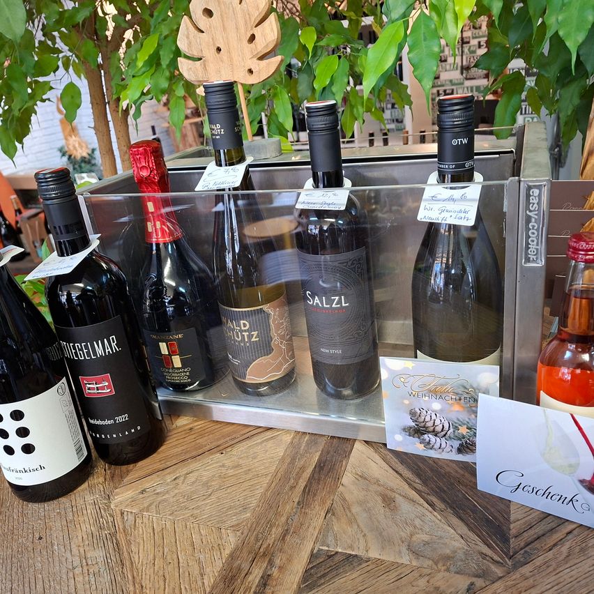 Ein Holztisch zeigt eine Glasbox mit sechs Weinflaschen. Die Flaschen sind mit Namen wie 'Salz' und 'Friegelmar' beschriftet. Eine Weihnachtskarte mit Tannenzapfen und einem 'Geschenk'-Text liegt auf dem Tisch.