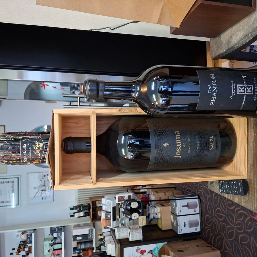 Zwei Weinflaschen in einer Holzkiste in einem Geschäft. Eine Flasche ist mit 'Das Phantom' und die andere mit 'Josanna' beschriftet. Das Geschäft hat viele Artikel zur Schau, darunter Weinflaschen, Kisten und eine Fernbedienung.