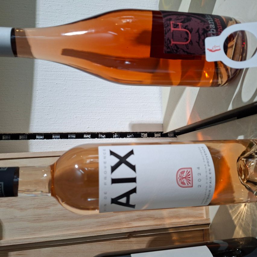 Zwei Flaschen Wein sind ausgestellt, eine mit dem Etikett 'AIX' und eine andere mit dem Etikett 'B', beide mit Korken und auf einem Holzregal.