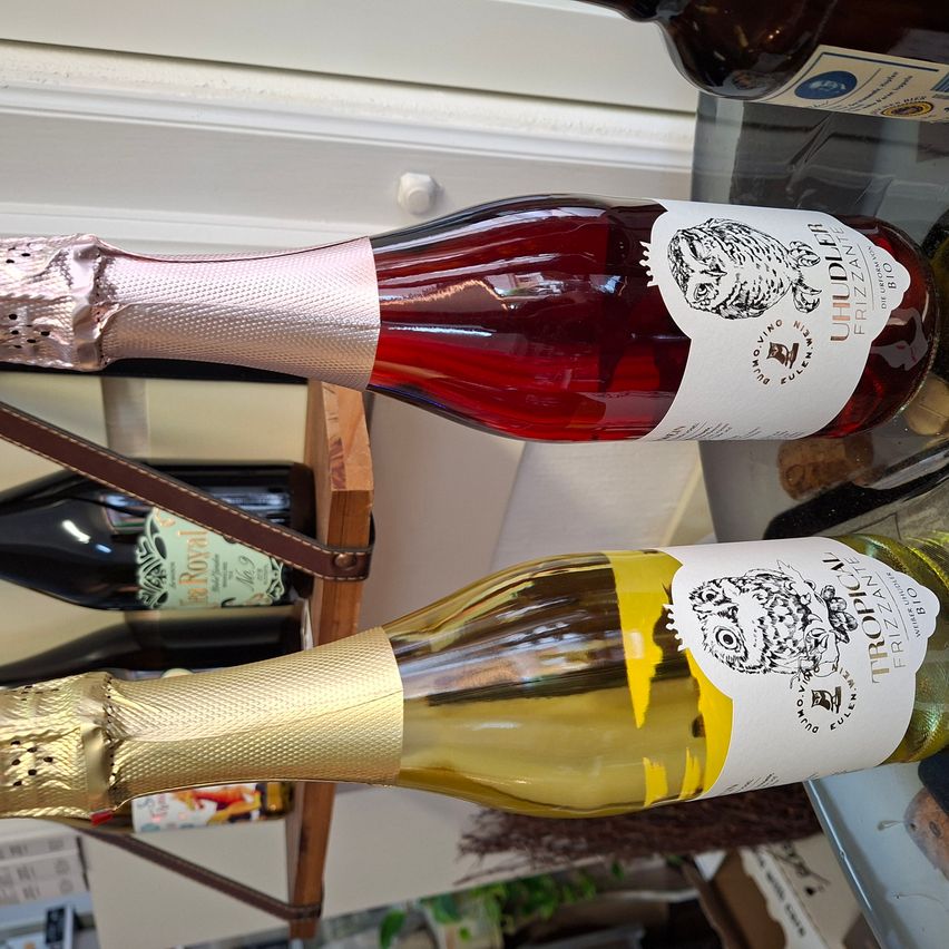 Zwei Flaschen Uhdeler Frizzante Wein, eine rote und eine weiße, sind auf einem Glastisch ausgestellt. Die Flaschen haben Etiketten mit Eulenillustrationen.