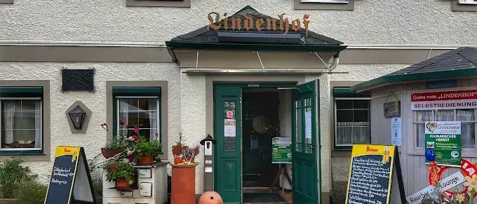 Die Vorderseite eines weißen Gebäudes mit einem Schild, das 'Lindenhof' liest. Draußen befinden sich Topfpflanzen und Blumen. Zwei Tafeln mit Schriftzügen und einer Tür sind sichtbar. Ein kleines Gebäude mit einem Schild, das 'Selbstbedienung' liest, befindet sich rechts.