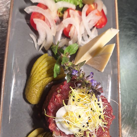 Ein Teller mit einem Steak, belegt mit einem rohen Ei, Sprossen und Blumen, begleitet von Gurken, Zwiebeln und Käse.