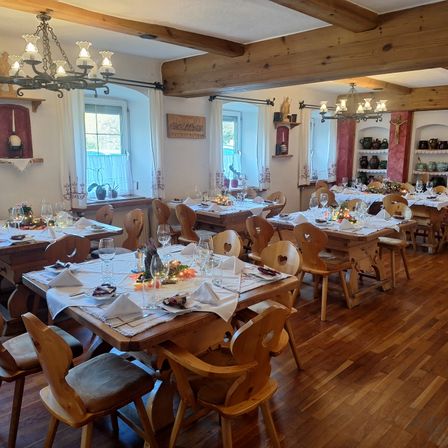 Ein leerer Speisesaal mit Holzboden und herzförmigen Stühlen. Die Tische sind mit Servietten, Weingläsern und Tellern gedeckt. Kronleuchter hängen von der Decke, und es gibt Fenster mit weißen Vorhängen.