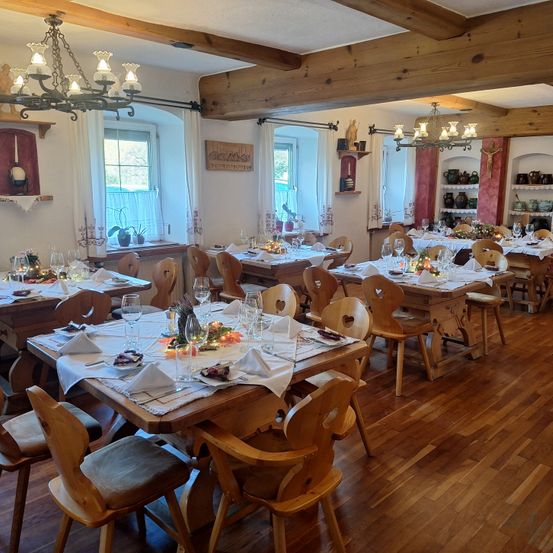 Ein leerer Speisesaal mit Holzboden und herzförmigen Stühlen. Die Tische sind mit Servietten, Weingläsern und Tellern gedeckt. Kronleuchter hängen von der Decke, und es gibt Fenster mit weißen Vorhängen.