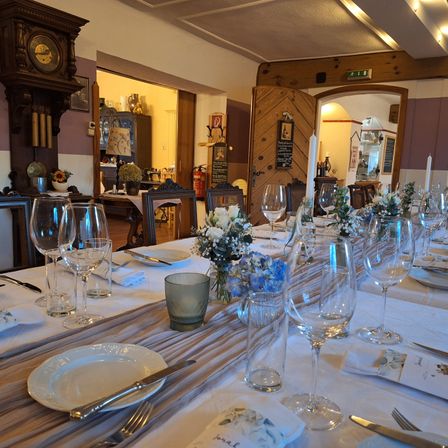 Ein langer Esstisch mit leeren Stühlen und Tellern, die mit Besteck und Weingläsern besetzt sind. Es gibt Blumen auf dem Tisch. Hinter dem Tisch steht eine Uhr und ein Schrank mit einer Lampe. In der Ferne ist eine offene Tür.