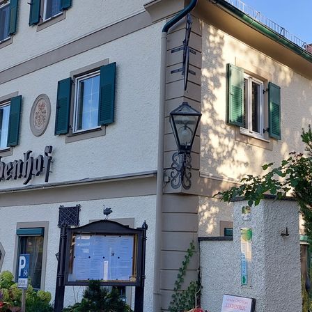 Ein Gebäude mit mehreren Fenstern und grünen Fensterläden heißt "Behrhof." Es hat ein Schild mit einem Menü, eine Lampe und eine Pflanze mit grünen Blättern.