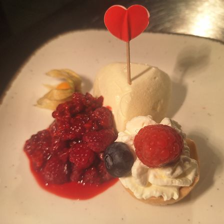 Ein Dessert auf einem weißen Teller mit einem herzförmigen roten Objekt, einer Kugel Eiscreme, einer Beerenkompottsauce, Schlagsahne und einer Himbeere obenauf.