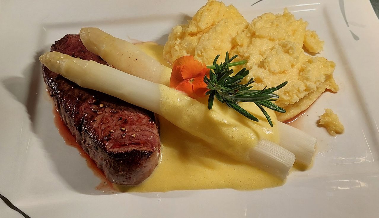 Ein Teller mit einem in Sauce getunkten Steak, Spargel und Kartoffelpüree mit Blumen- und Rosmaringarnitur.