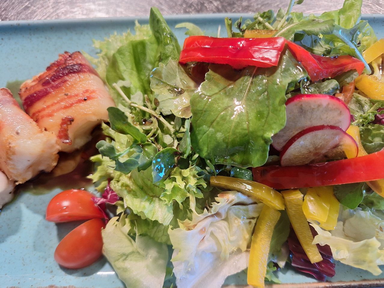 Ein Salat auf einem blauen Teller besteht aus gemischtem Blattsalat, Kirschtomaten, roten Paprikastreifen und einem Stück gegrilltem Fleisch.