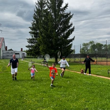 Eine Gruppe von Menschen, einschließlich Erwachsener und Kindern, rennt und spielt Tennis auf einem Rasenfeld. Ein Baum und ein Gebäude sind im Hintergrund.