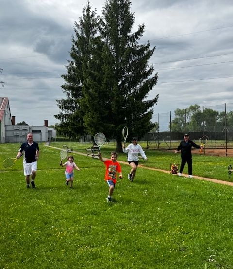 Eine Gruppe von Menschen, einschließlich Erwachsener und Kindern, rennt und spielt Tennis auf einem Rasenfeld. Ein Baum und ein Gebäude sind im Hintergrund.