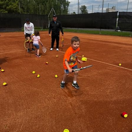 Ein junger Junge übt Tennis auf einem Platz mit mehreren verstreuten Tennisbällen. Ein Erwachsener und ein Kind beobachten in der Nähe.
