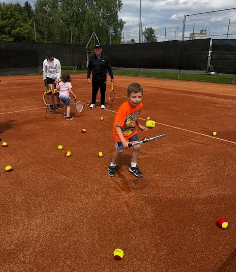 Ein junger Junge übt Tennis auf einem Platz mit mehreren verstreuten Tennisbällen. Ein Erwachsener und ein Kind beobachten in der Nähe.