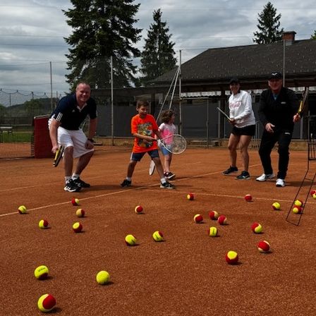 Eine Gruppe von Tennisspielern übt auf einem Platz mit vielen Tennisbällen. Zwei Männer stehen in der Nähe des Netzes, während ein Junge und ein Mädchen üben.