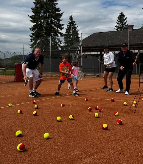 Eine Gruppe von Tennisspielern übt auf einem Platz mit vielen Tennisbällen. Zwei Männer stehen in der Nähe des Netzes, während ein Junge und ein Mädchen üben.
