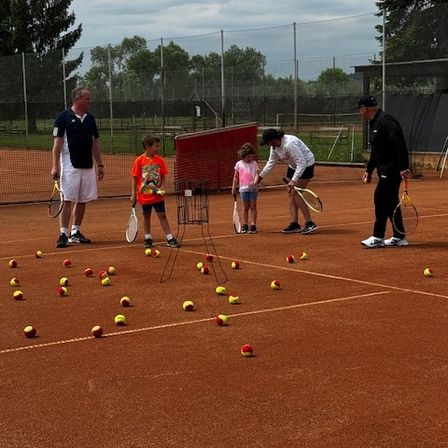 Mehrere Personen, darunter Erwachsene und Kinder, sind auf einem Tennisplatz und halten Schläger. Eine Frau bringt einem jungen Mädchen das Spielen bei. Tennisbälle sind auf dem Platz verstreut.