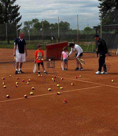 Mehrere Personen, darunter Erwachsene und Kinder, sind auf einem Tennisplatz und halten Schläger. Eine Frau bringt einem jungen Mädchen das Spielen bei. Tennisbälle sind auf dem Platz verstreut.