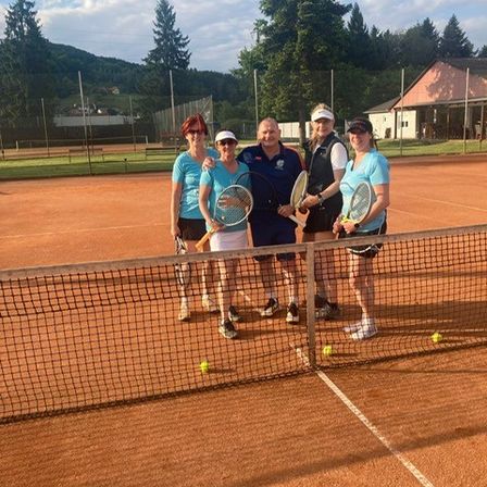 Fünf Personen stehen auf einem Tennisplatz und halten alle Tennisschläger in den Händen. Sie posieren für ein Foto, mit Tennisbällen um sie herum. Bäume und ein Gebäude sind im Hintergrund zu sehen.