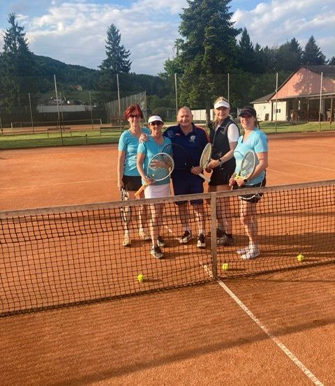 Fünf Personen stehen auf einem Tennisplatz und halten alle Tennisschläger in den Händen. Sie posieren für ein Foto, mit Tennisbällen um sie herum. Bäume und ein Gebäude sind im Hintergrund zu sehen.