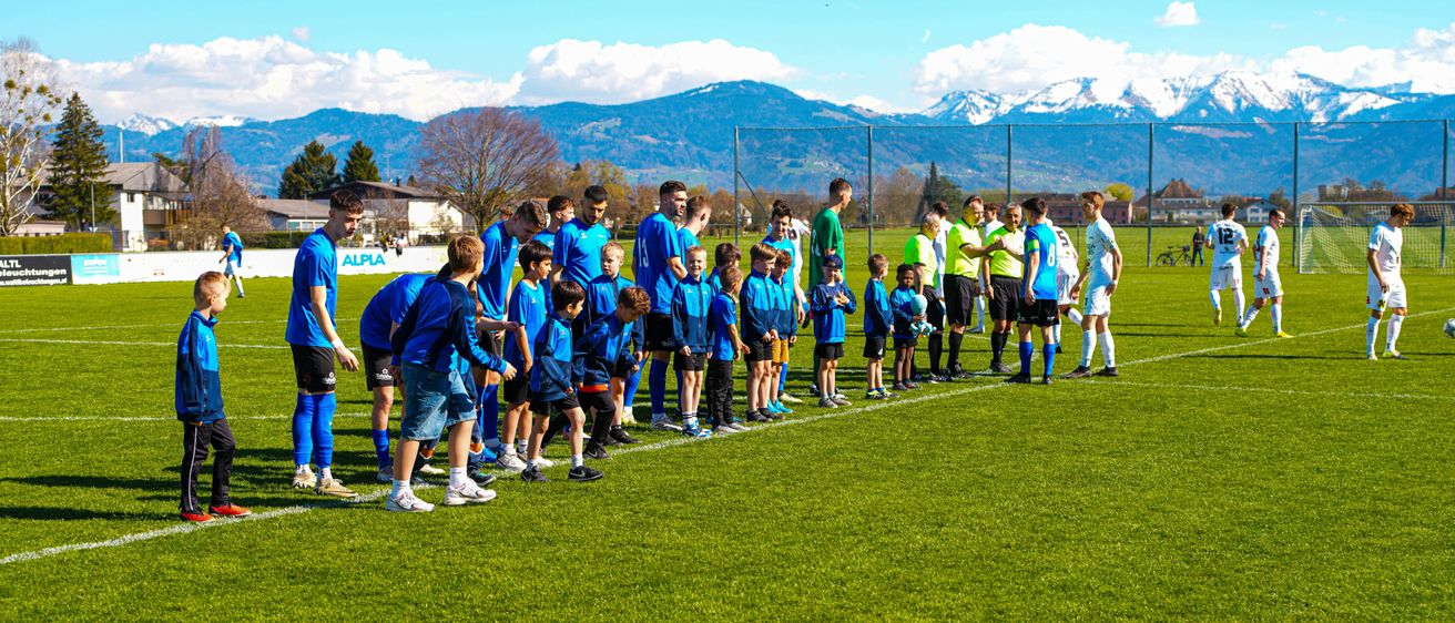 Eine Gruppe junger Fußballspieler in blauen Uniformen steht auf einem Feld mit Erwachsenen und Bergen im Hintergrund.
