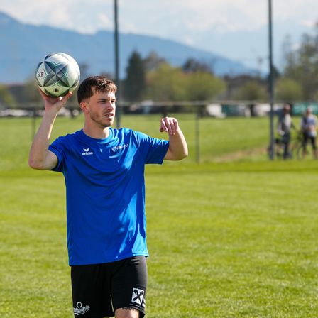 Ein Mann in einem blauen T-Shirt hält einen Fußball in der Luft auf einem grünen Feld mit Bergen im Hintergrund.