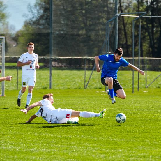 Drei Fußballspieler sind auf einem Spielfeld. Ein Spieler liegt auf dem Boden, während ein anderer Spieler springt, um den Ball zu treten. Der dritte Spieler steht in der Nähe des Tores.