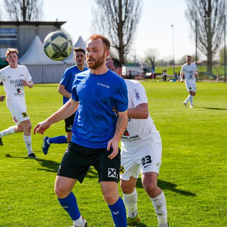 Ein Fußballspieler in einem blauen Trikot versucht, den Ball zu köpfen, während zwei Gegner in weißen Trikots versuchen, ihn zu blockieren, auf einem Rasenfeld mit Bäumen im Hintergrund.