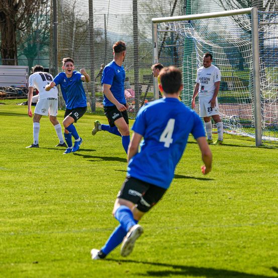 Fußballspieler in blauen und weißen Trikots spielen auf einem Feld. Ein Spieler in Blau läuft auf das Tor zu, während andere jubeln. Ein Spieler in Weiß beobachtet von hinter dem Tor.