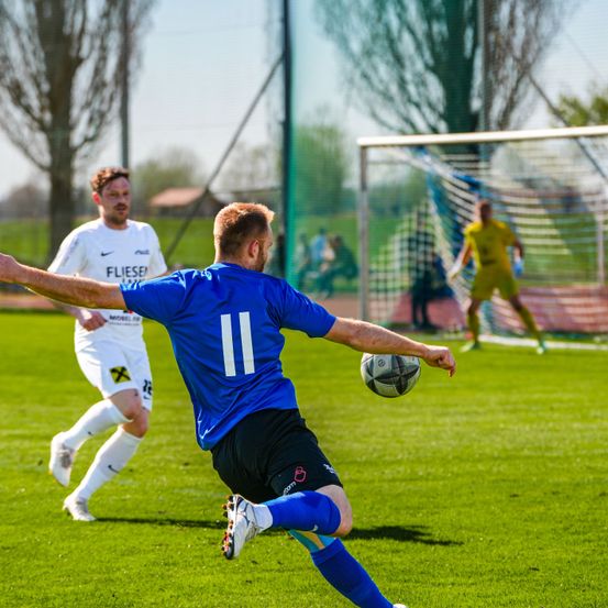 Zwei Fußballspieler sind auf einem Feld. Einer trägt ein blaues Trikot mit der Nummer 11 und tritt einen Ball. Der andere ist in einem weißen Trikot und läuft.