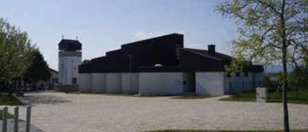 Ein modernes Gebäude mit schwarzem Dach und weißen Wänden. Ein Turm steht neben ihm. Das Gebäude ist von Gras und Bäumen umgeben.