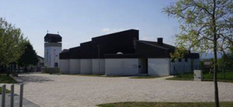 Ein modernes Gebäude mit Turm steht auf einem Feld, umgeben von Gras und Bäumen unter klarem Himmel.