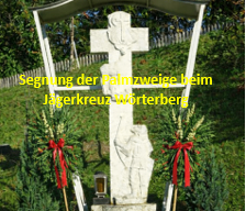 Ein Kreuz mit einer geschnitzten Figur steht auf einer Rasenfläche, umgeben von roten Schleifen und grünen Pflanzen. Der Text lautet 'Segnung der Palmzweige beim Jägerkreuz-Wörtenberg'. Hinter dem Kreuz befindet sich ein Holzzaun und Bäume.