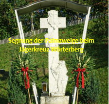 Ein Kreuz mit einer geschnitzten Figur steht auf einer Rasenfläche, umgeben von roten Schleifen und grünen Pflanzen. Der Text lautet 'Segnung der Palmzweige beim Jägerkreuz-Wörtenberg'. Hinter dem Kreuz befindet sich ein Holzzaun und Bäume.