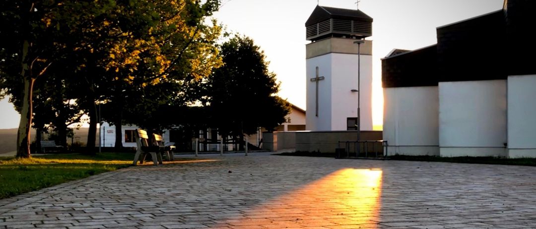 Ein Kirchturm mit einem Kreuz, umgeben von Bäumen und einer Bank im Vordergrund, wobei das Sonnenlicht Schatten auf den Boden wirft.