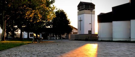 A church tower with a cross on the side, surrounded by trees, with a paved area in the foreground.