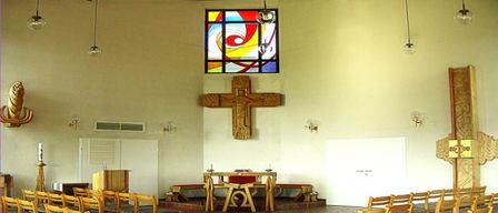 Ein leerer Kircheninnenraum mit Holzbänken, einem hölzernen Altar und einem Buntglasfenster mit einem Kreuz darüber. Die Wände sind weiß, und es gibt brennende Kerzen und Lampen.