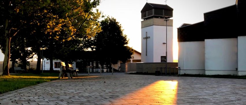 Ein Kirchturm mit einem Kreuz steht in einer ruhigen Außenumgebung, umgeben von Bäumen und einem gepflasterten Weg.
