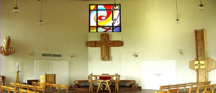 An empty church interior with a wooden cross on the wall, a stained glass window, and wooden pews.