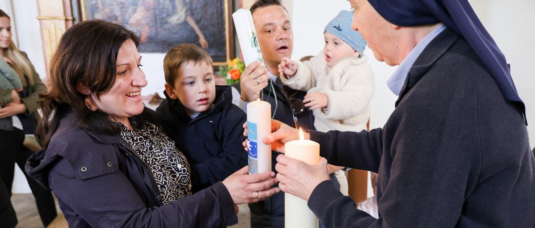 Eine Familie und eine Nonne halten Kerzen, wahrscheinlich in einer Kirche. Die Familie besteht aus zwei Kindern, einem Baby und einem erwachsenen Mann und Frau.
