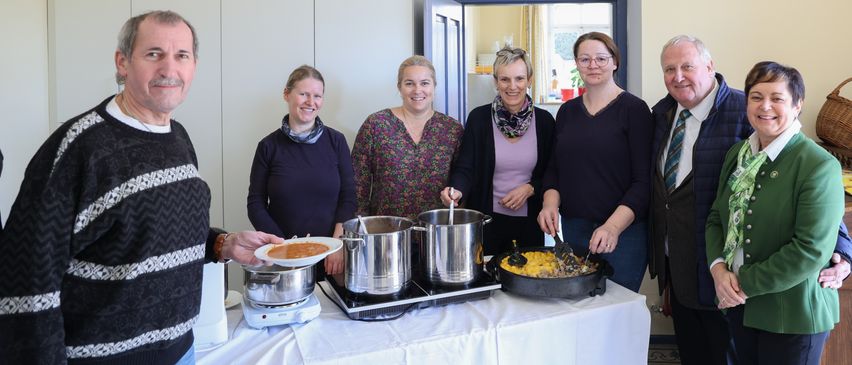 Sechs Personen stehen in einem Raum und kochen mit Töpfen und Pfannen auf einem Herd. Eine Person hält einen Teller mit Essen.