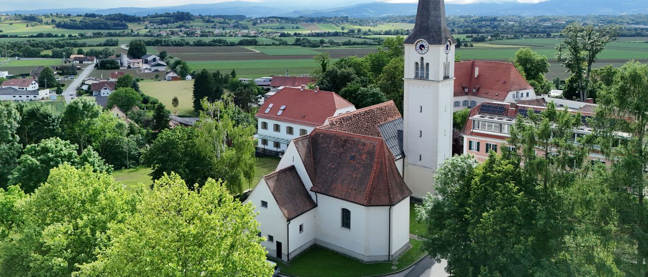 Luftaufnahme einer Kirche mit einem Kirchturm und einem roten Dach, umgeben von Bäumen, Häusern und grünen Feldern unter einem bewölkten Himmel.