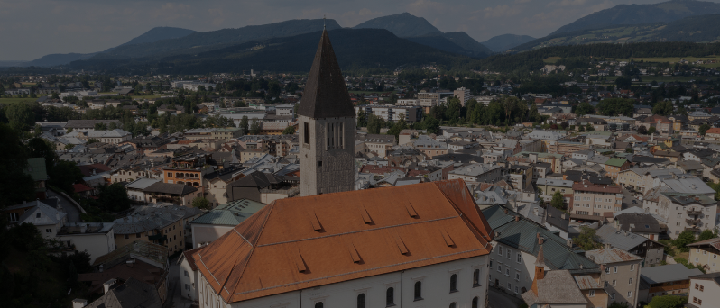 Luftaufnahme von Pfarrkirche Hallein, einer Kirche mit hohem Turm und rotem Dach in einer Stadt mit vielen Gebäuden, umgeben von Bergen.