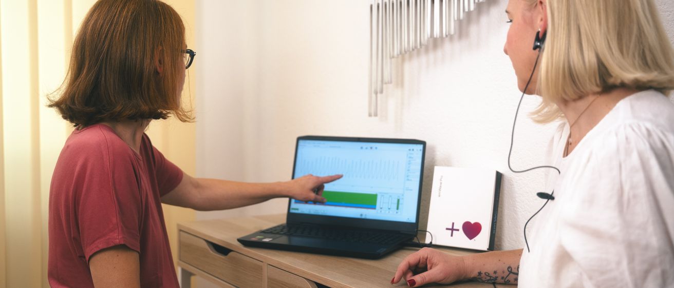 Eine Frau zeigt mit dem Finger auf einen Laptop-Bildschirm mit einem Diagramm, während eine andere Frau zusieht. An der Wand befindet sich eine Ausstellung von Stimmgabeln und eine weiße Box mit einem roten Herz und einem Pluszeichen.