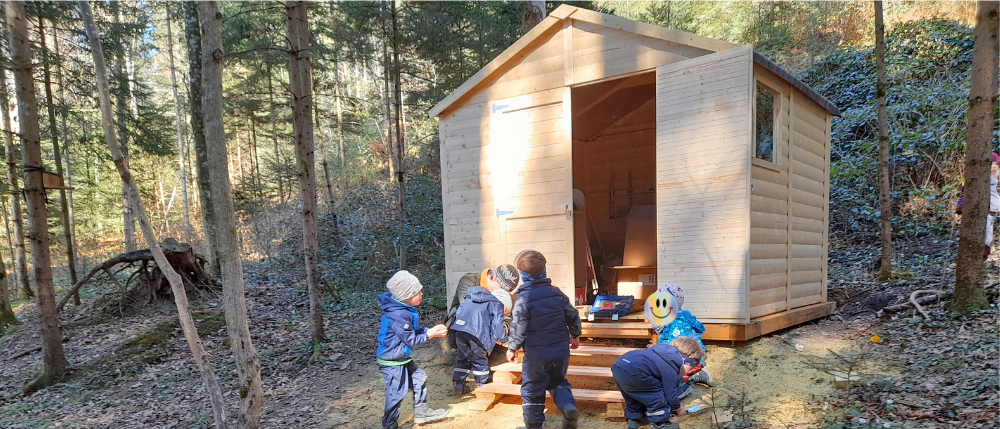 Mehrere Kinder in blauen Jacken arbeiten an einem Holzschuppen im Wald. Ein Kind trägt einen Hut.