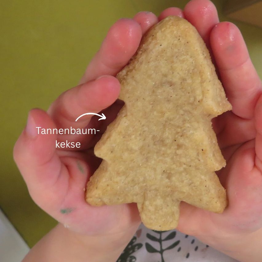 Eine Hand hält einen goldenen, baumförmigen Keks. Ein weißer Pfeil zeigt auf den Keks, mit dem Text 'Tannenbaum-kekse' darüber.